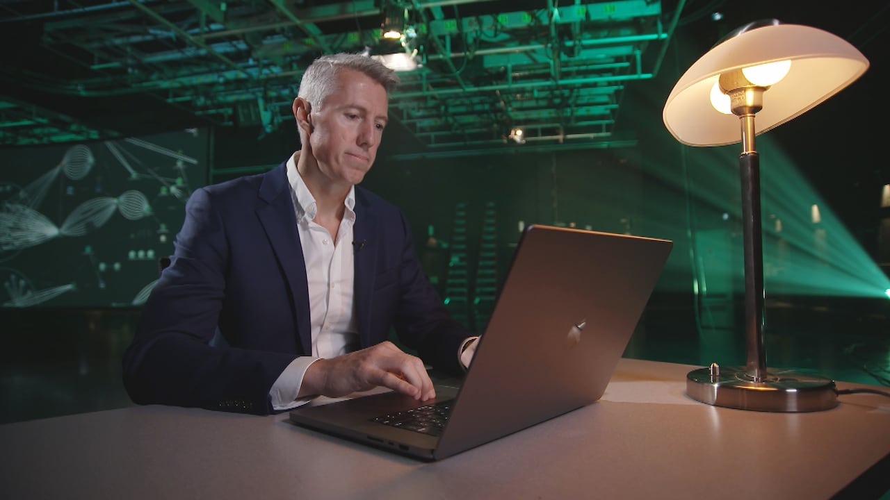 Kyle Armstrong sits at a desk typing on a laptop with a map of cryptocurrency transactions in the background. 