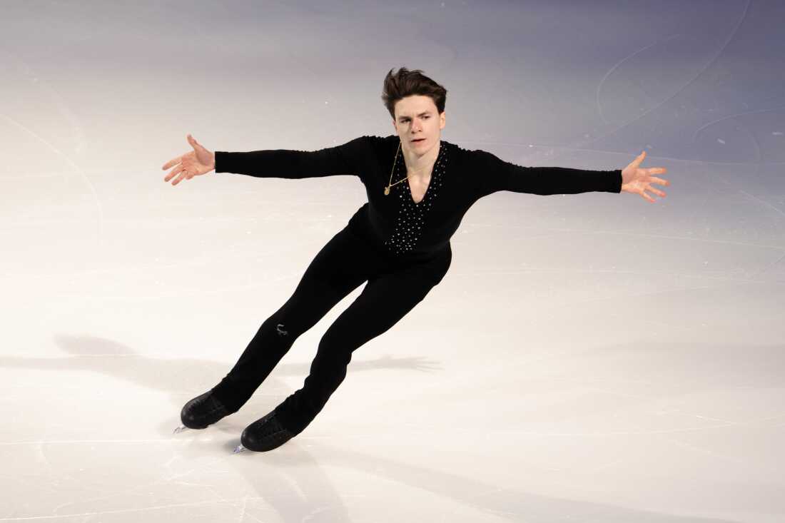 Figure skater Maxim Naumov performs in exhibition on ice. He has arms outstretched and is wearing an all black costume with rhinestones on the chest.