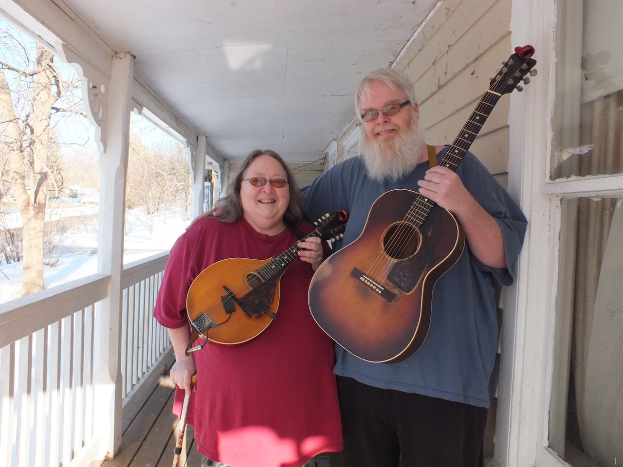 a woman with glasses and a red shirt hold a small guitar next to a taller man with a blue shirt and beard holding a guitar