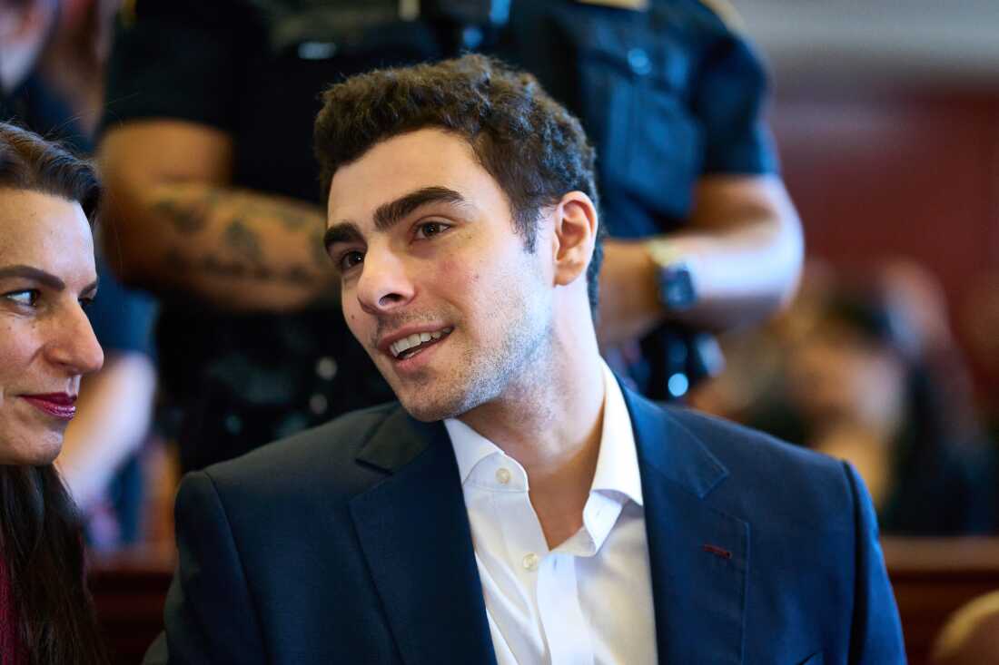 Luigi Mangione wearing a white collared shirt and navy blue suit blazer, smiles as he leans over to speak with his attorney in Manhattan Criminal Court.