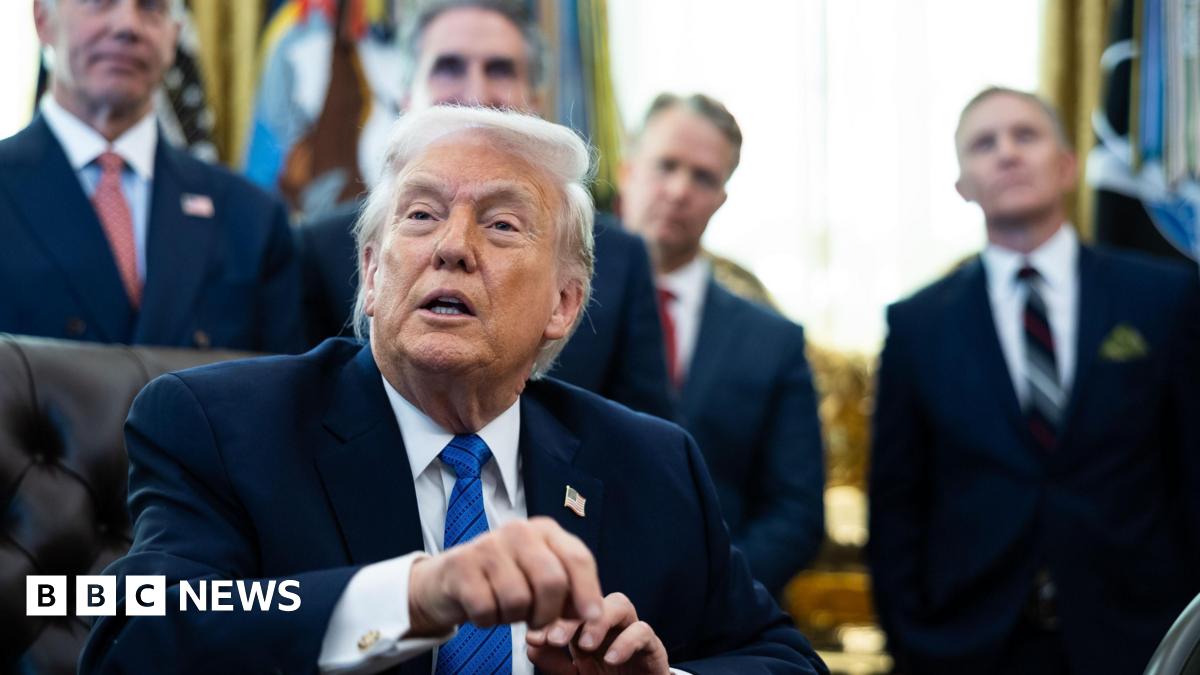Donald Trump pictured sat behind his desk in the Oval Office. He wears a black suit jacket, white shirt, blue tie, and a pin of the US flag. Behind him stand four male officials in suits.