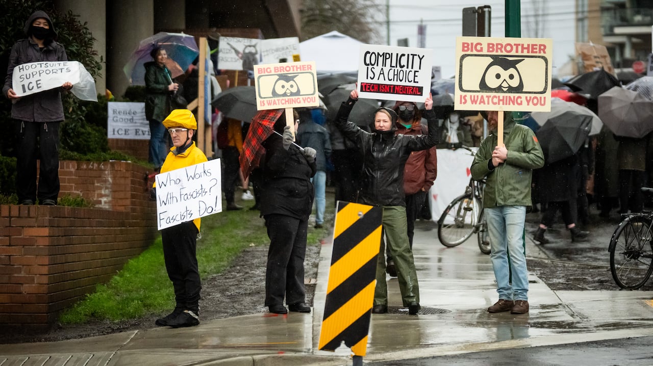 A group of people hold up signs in rainy conditions criticizing Hootsuite, complicity, and saying only fascists work with fascists.