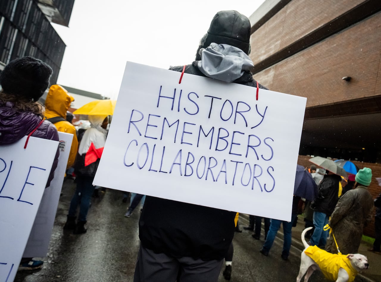 A person holds a sign behind them reading 'History remembers collaborators'.