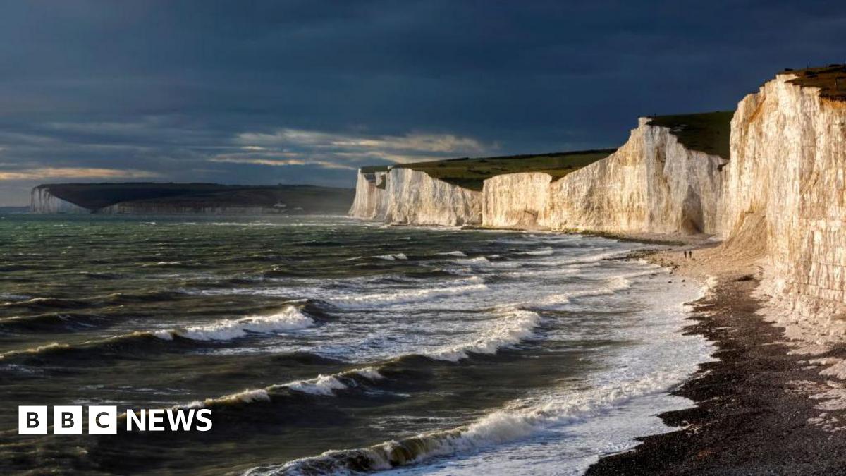 Waves crash to the shore beside some large white cliffs. The sky looks dark and stormy.