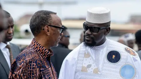Guinea Presidency A side view of Paul Kagame in a patterned African shirt talking to Mamady Doumbouya in a white boubou, white hat and dark glasses.