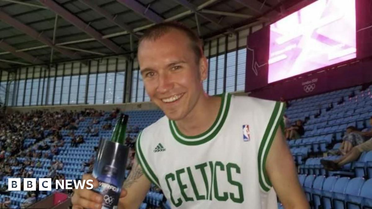 Sean Fitzgerald at a sports stadium, wearing a Celtics T-shirt and holding a drink. He is smiling and looking happy. There are people in the blue seats behind him.