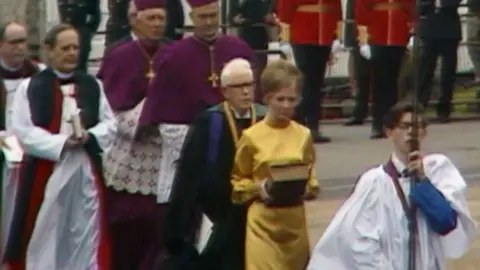 Cwmni Da/S4C A young Kenner Jones carries a pole with an out of sight cross at the top of it. He is wearing choirboy clothing and glasses. He is leading a procession with a woman, three priests and two bishops.
