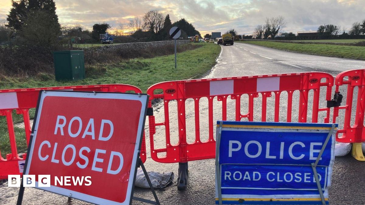 Road closure signs in front of red barriers that have been put up on Cirencester Road. Emergency service vehicles can be seen in the distance.