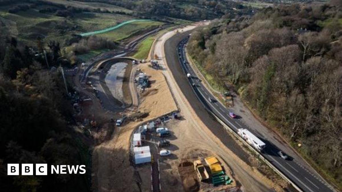 An aerial view of a new stretch of road, which is still under construction, beside an existing road being used by traffic up a hill between two woodlands.