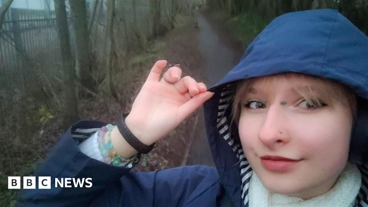 A selfie image of Amelka from the shoulders up, she is wearing a navy rain coat with the hood up and a white scarf. She is on a gloomy walking path, it seems to have been raining and it's cold.