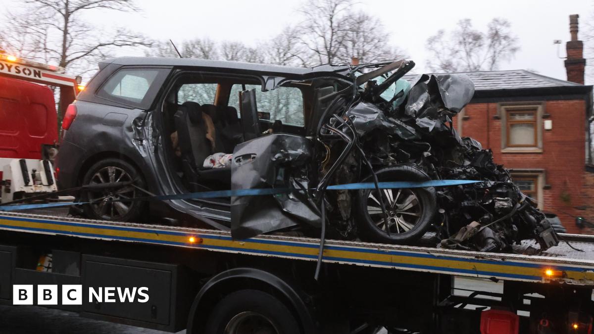 A dark Seat car with its front bonnet completely smashed and both right doors torn off is towed away on the back of a lorry past houses.