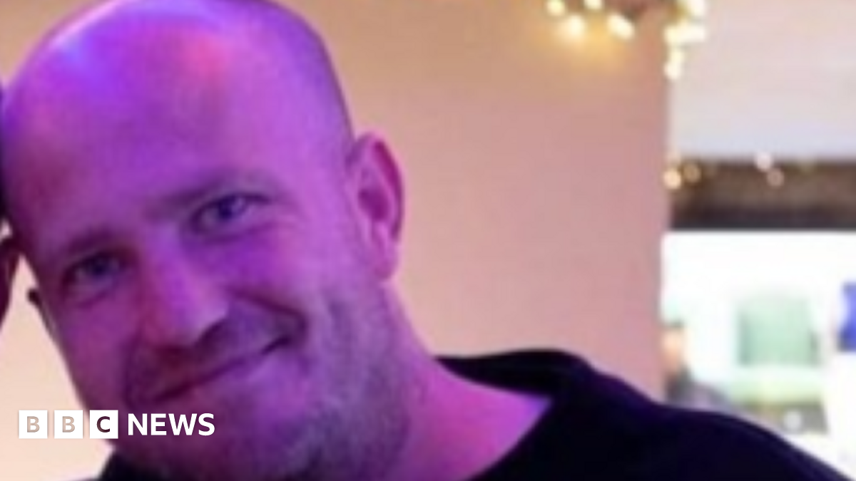Brian Russell, a shaven headed man wearing a black top, smiles for the camera while standing indoors.