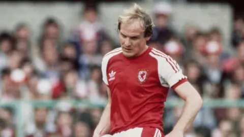 Getty Images Terry Yorath pictured in a Wales kit in 1980. He is looking at a football as he kicks it. The crowd in the stadium can be seen blurred in the background.