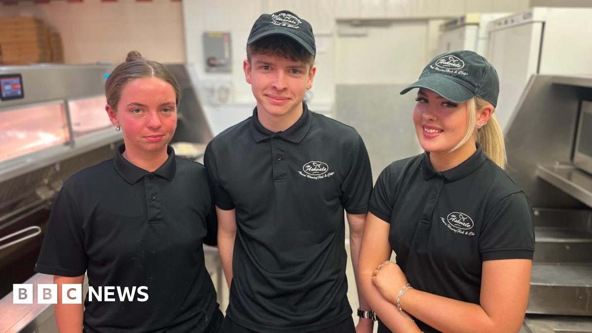 Two young women and one young man (centre) in their chip shop uniforms of a black polo shirt and trousers and a skip cap smile for the camera. They are standing beside the fat fryers and food prep area, all in stainless steel.