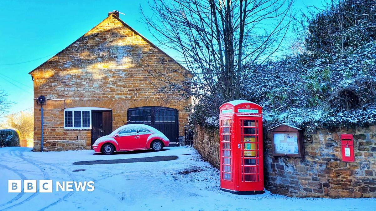 A sandstone how with a brown wood door and brown wooden garage doors. In front of it is a red VW Beatle car. to the right is a stone wall, red phone box and red post box embedded in the wall. The wall is in front of a bank with bushes and tree. The ground and foliage is covered in a light dusting of snow.