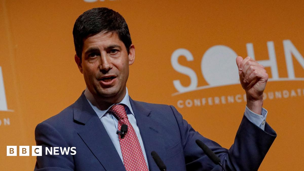 Kevin Warsh wearing a blue suit, light blue shirt and red patterned tie speaking at a conference in front of an orange background