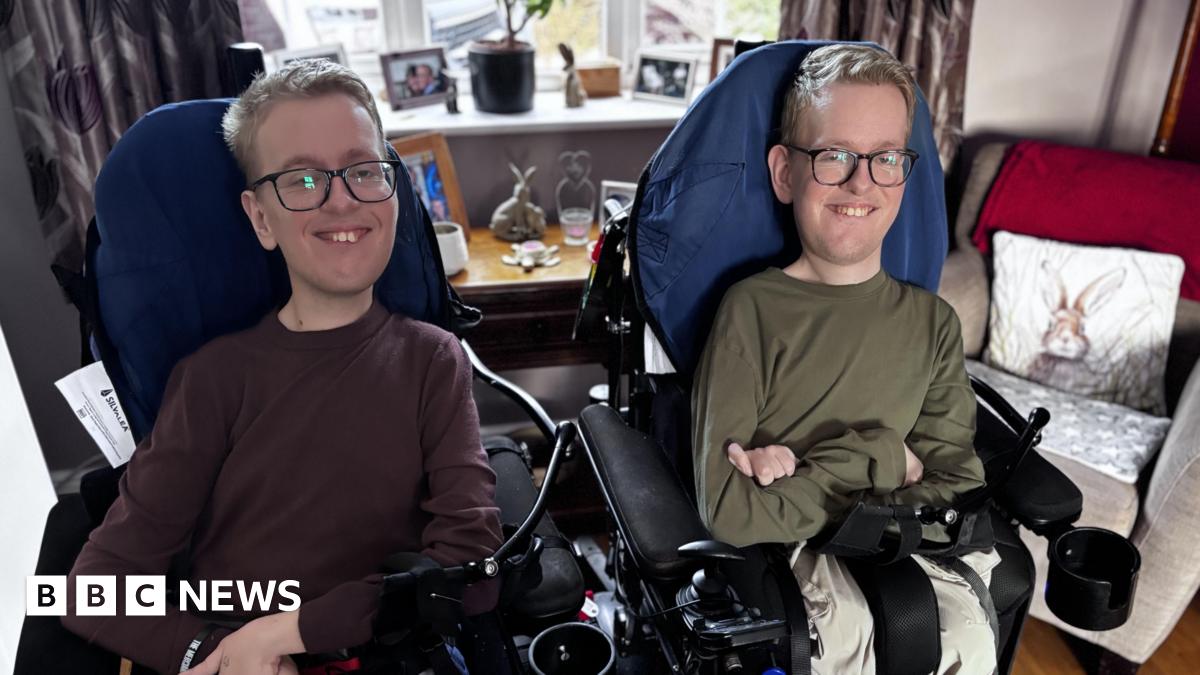Identical twins Sam and Alex Bolton pictured sat in their wheelchairs in their living room smiling.