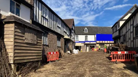 Sharon Amery A street with black and white buildings that is part of a film set. There is a large blue board against one building at the end of the street, and brown wooden wagons next to houses. There is straw lining the floor