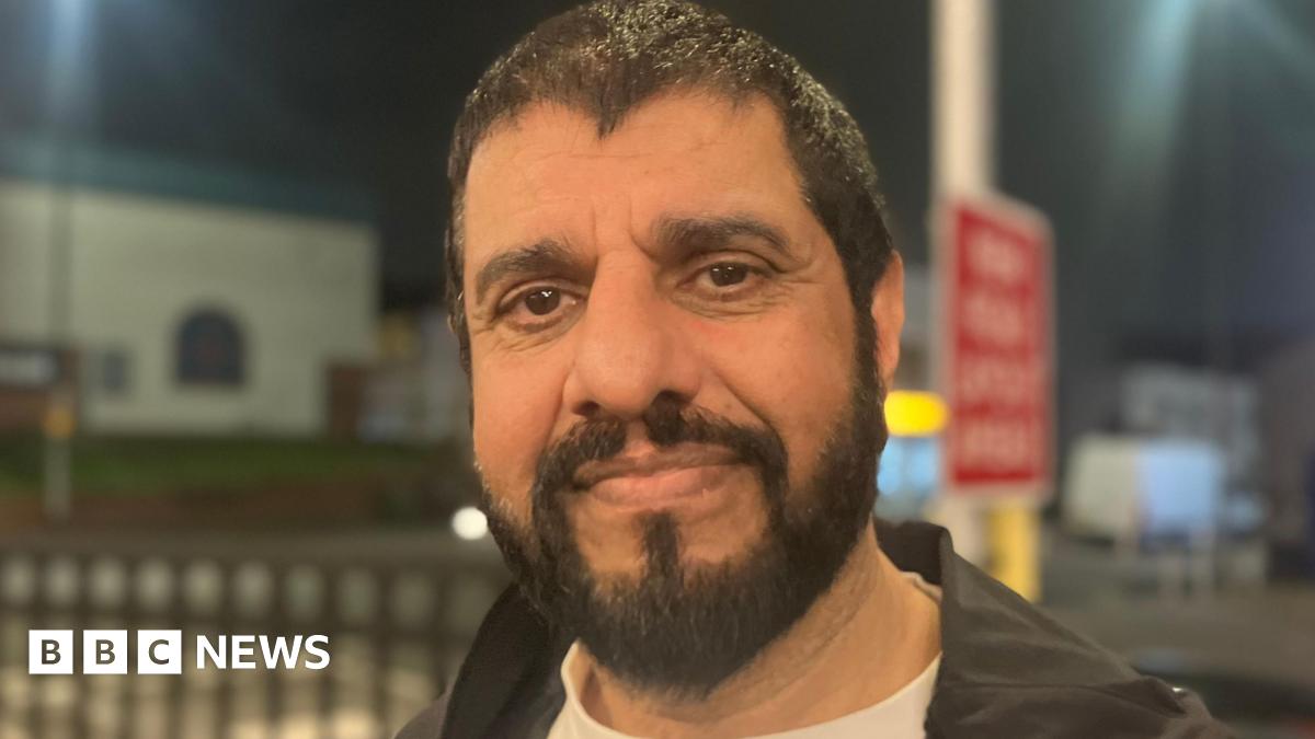 A man with a beard smiles in the dark. Road signs are illuminated behind him.
