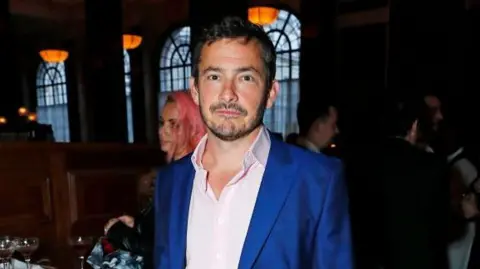 Getty Images Giles Coren attends the press night performance of "Opening Night" at The Gielgud Theatre on March 26, 2024 in London, England. He wears a blue suit jacket and a pink shirt. It is a head and shoulders shot.