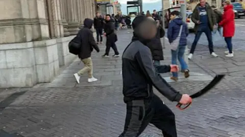 Alan Simpson Photography Person holding a large bladed weapon walking across a city centre street, with pedestrians nearby and buildings lining the pavement.