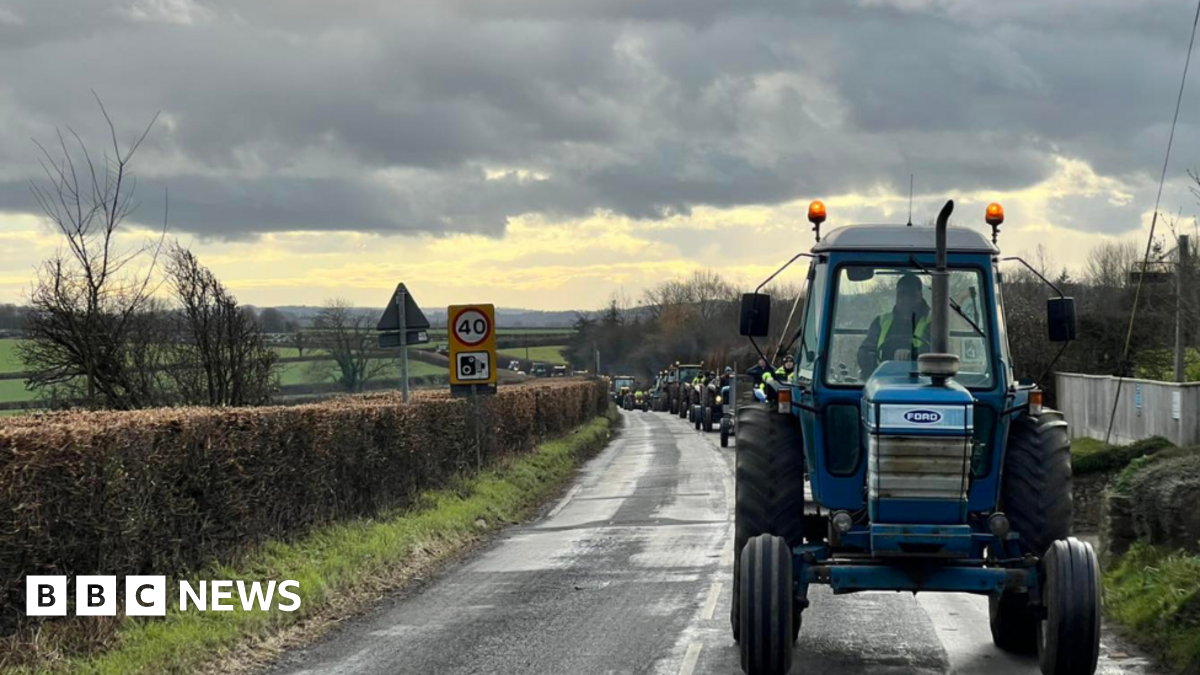 A line of tractors driving down a country road beneath a cloudy sky. Houses are visible on the right of the image while a hedge and fields can be seen on the left.