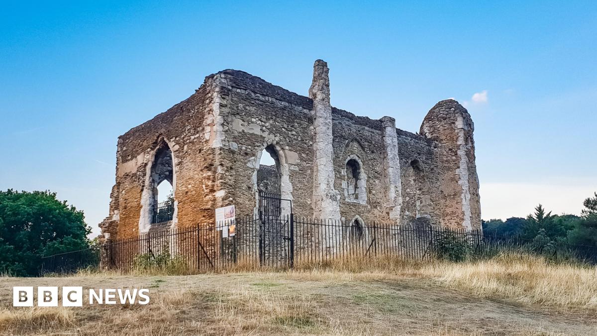 A ruined stone building without a roof or windows. It is surronded by yellow grass and behind a black metal fence.