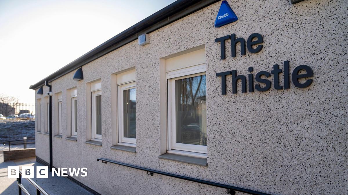 The entrance to a brick building on a sunny day. A sign saying The Thistle is hanging on a wall.