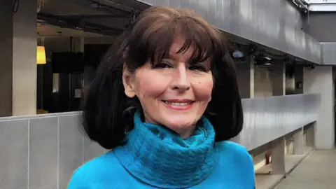 Marquis wearing a bright blue textured turtleneck sweater, standing outdoors in a modern urban setting with gray walls and glass panels in the background.