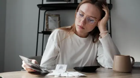 Getty Images Woman, with her head resting on her hand, looks at receipts while sitting at a table with a teacup and calculator in front of her.