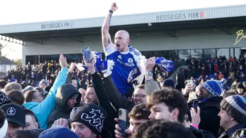 PA Media Macclesfield's Josh Kay raises his arm in the air celebrating while surrounded by fans on the pitch. His mouth is open and he looks to be cheering. He appears to be on the shoulders of a team-mate and raised above the surrounding fans.