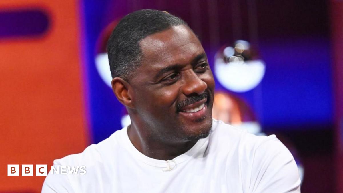 Sir Idris Elba looks away from the camera in a white shirt and smiles. The background is blurred.
