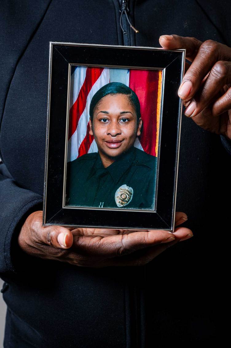 A woman holds a framed portrait of her daughter, depicting her wearing a police uniform in front of an American flag.