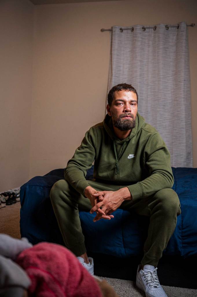 A man wearing a green sweatsuit sits on the edge of a bed.