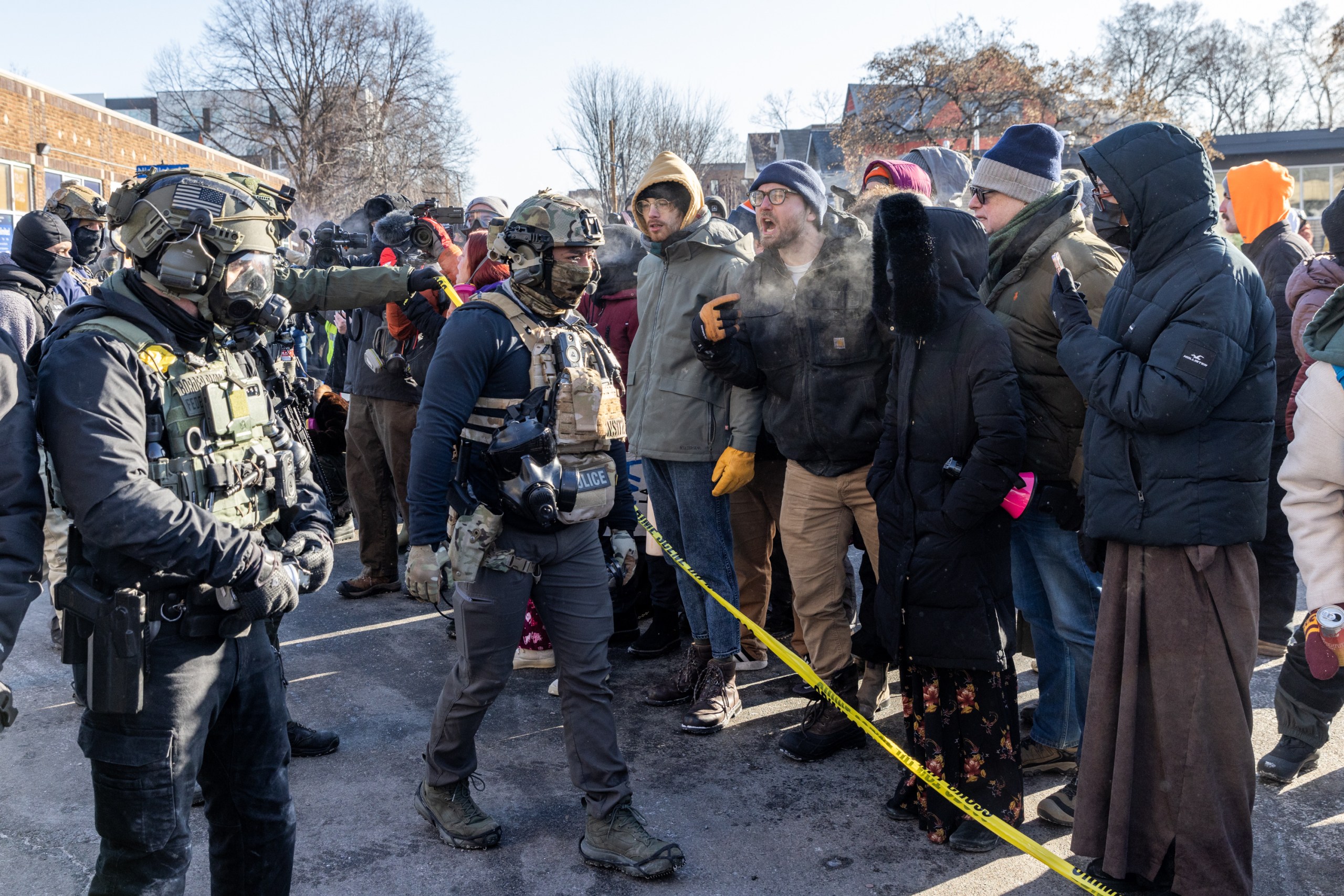 Several men in tactical vests and helmets have their backs to the camera, facing a row of people in street clothes. Members of the crowd are shouting and their breath is visible. A strand of yellow police tape cuts across the scene.
