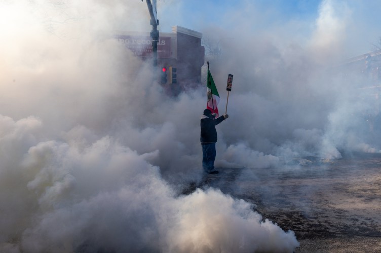 A man holds a Mexican flag and a protest sign in a cloud of tear gas. A street light is just visible behind him, but otherwise the cloud of gas is so thick and tall that the rest of the street is obscured.