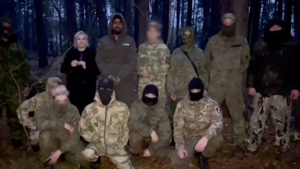 Azarnykh in a dark coat standing next to nine people in military clothing, with dense forest in the background. Next to her is Habib, who is wearing a khaki-coloured hood but his face is visible. The other people's faces are covered with black balaclavas or scarves, except for one whose face has been blurred. 