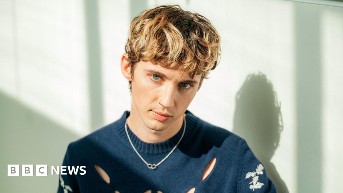 A portrait of Troye Sivan where he is looking at the camera with brownish hair. He has a nose ring on, a blue jumper and a necklace. He's infront of a white wall.
