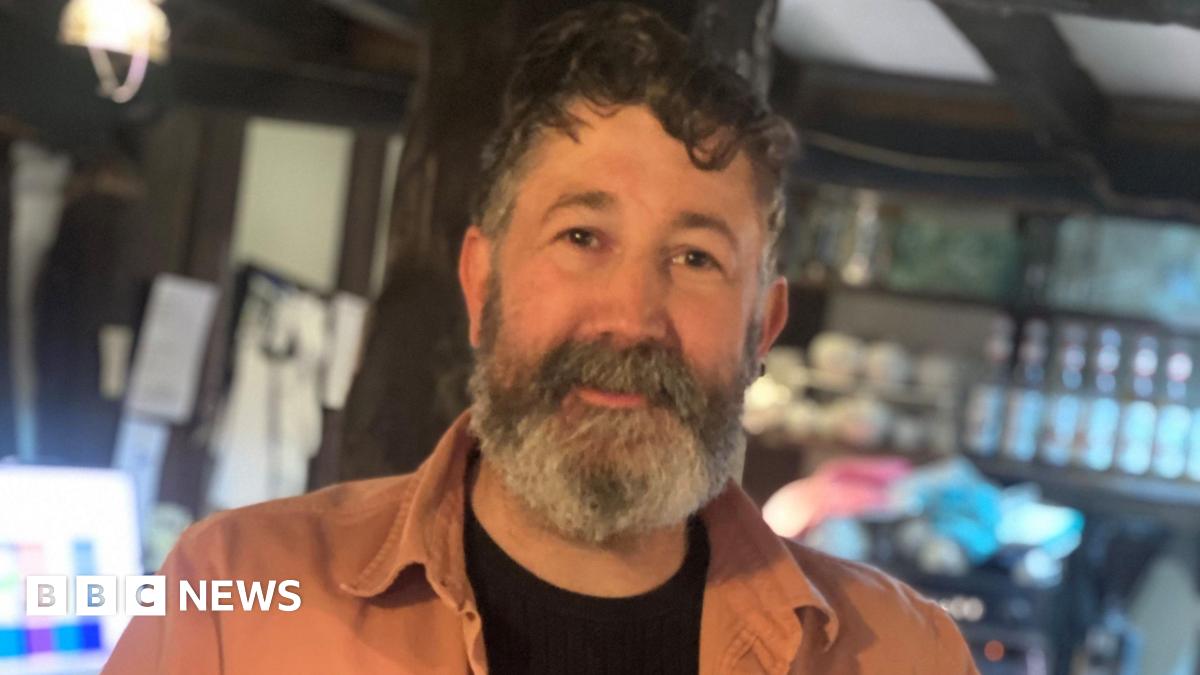 A middle-aged man with curly black hair and a grey beard. He is looking into the camera and is wearing a black t-shirt with orange shirt over the top. He is stood in a room with bottles and cups on shelving behind him, with a low beamed ceiling.
