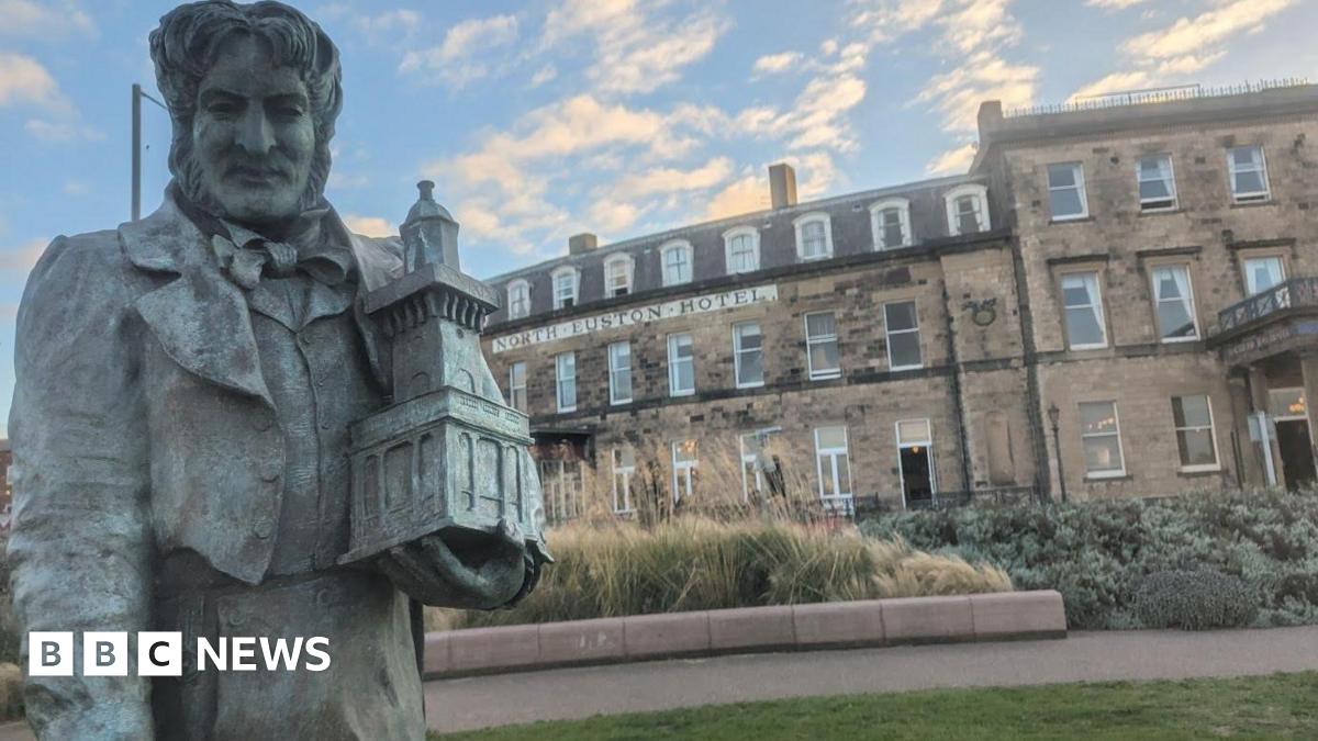 A veterinarian wearing blue scrubs and a stethoscope gently holds a young black and tan puppy, possibly a German Shepherd, in a clinical setting. The vet's gloved hand supports the puppy, conveying care and professionalism against a clean, light blue background.