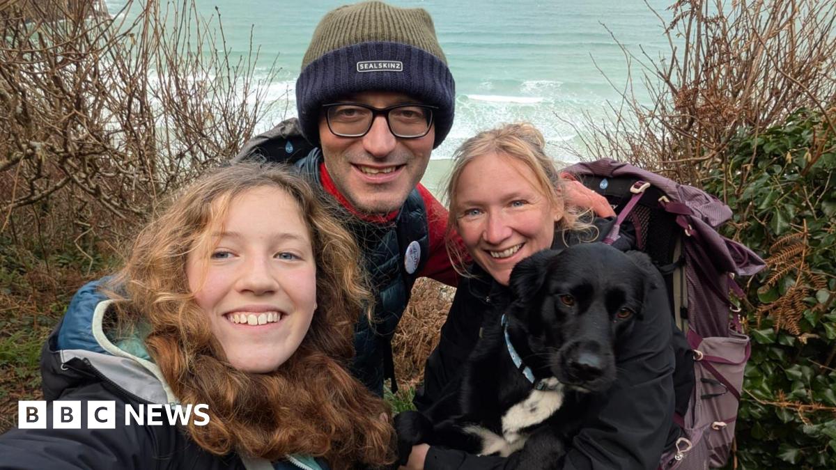 The family huddle together and smile at the camera. They are in outdoor walking gear and the sea is in the background. Tara, the mother, has the family's black and white dog on her lap.