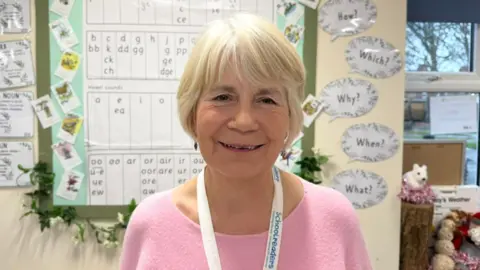 BBC / Grace McGrory The photo shows a woman smiling at the camera. She has short blonde hair and is wearing a pale pink jumper. She has a lanyard round her neck which reads 'Schoolreaders.' She's standing in a classroom, in front of a display which has lots of letters on it. There are also speech bubbles that have been stuck on the walls with various words written on them. 