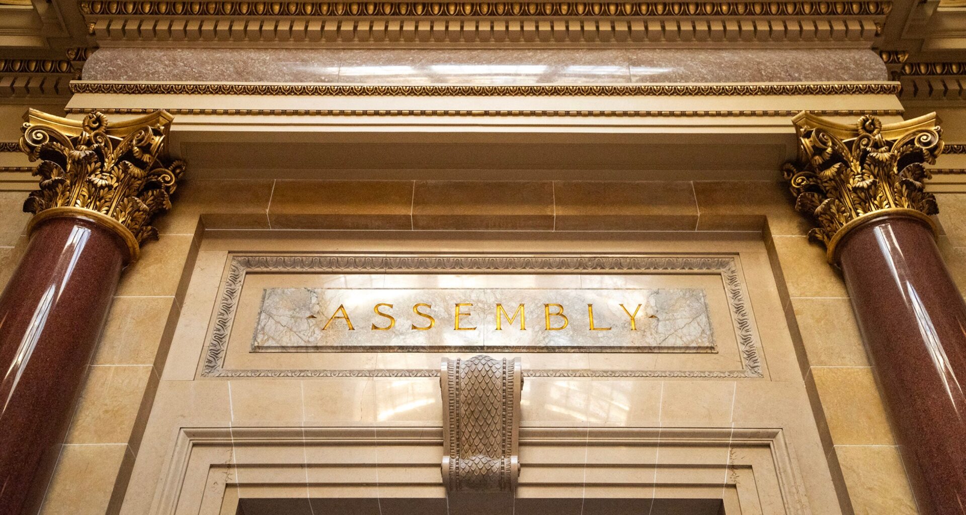 Ornate architectural detail above a doorway with the word ASSEMBLY engraved in gold letters, flanked by marble columns and decorative gold capitals.