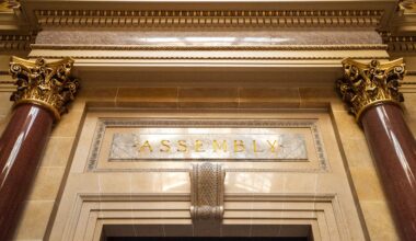 Ornate architectural detail above a doorway with the word ASSEMBLY engraved in gold letters, flanked by marble columns and decorative gold capitals.