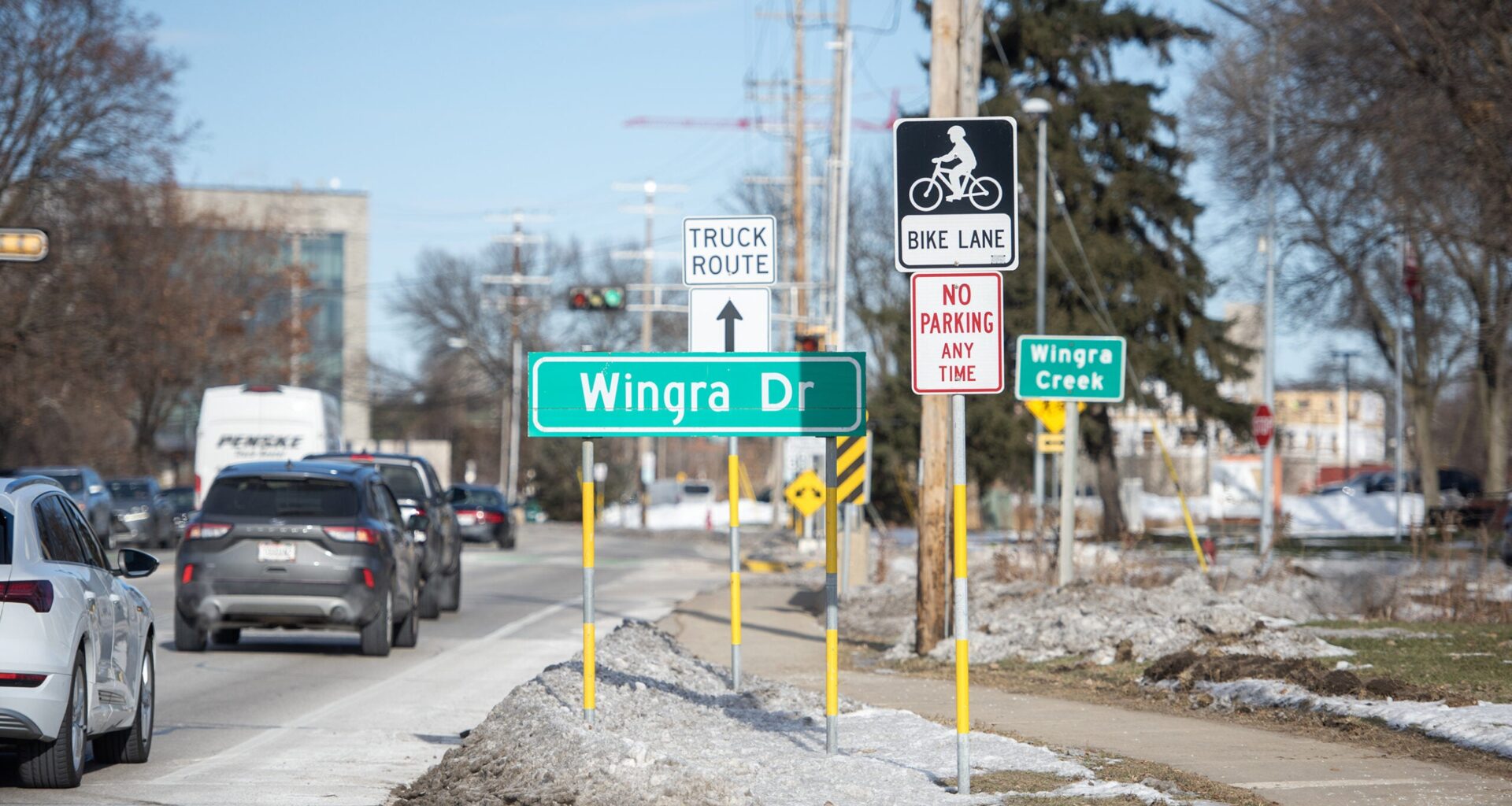 Cars drive past street signs for Wingra Dr and Wingra Creek, along with truck route and bike lane signs, on a city road with patches of snow.