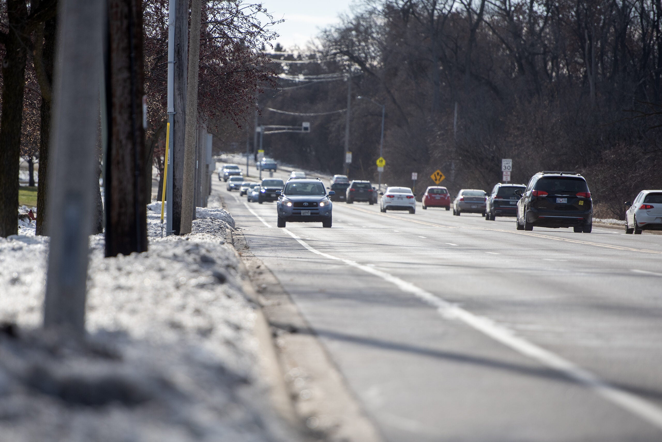 Cars drive along a multi-lane road bordered by snow-covered sidewalks and trees on a partly cloudy day.