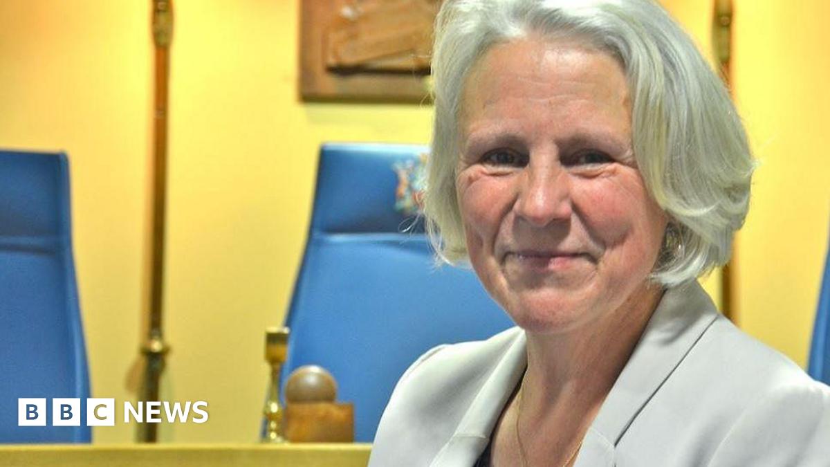 Rossendale Council leader Alyson Barnes sitting in council chambers with grey bobbed hair wearing a light grey suit jacket. She is smiling.