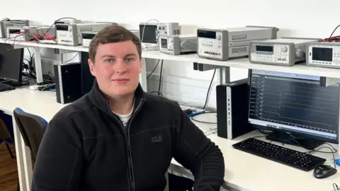 Alex Bethell Wearing a black fleece, Alex Bethell sits facing the camera with a computer and a shelf of electronic equipment behind him.