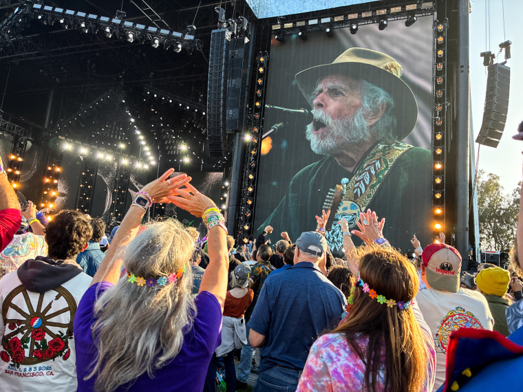 Bob Weir can be seen on a large screen next to a stage he performs on, in front of a large crowd outdoors