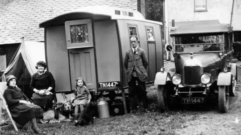National Motor Museum/Heritage Images/Getty Images A black and white photo of a family - two women, a man, a child and two dogs - in 1920s style clothing. They sit outside a 1926 Eccles model caravan which is set next to their car and a tent. 
